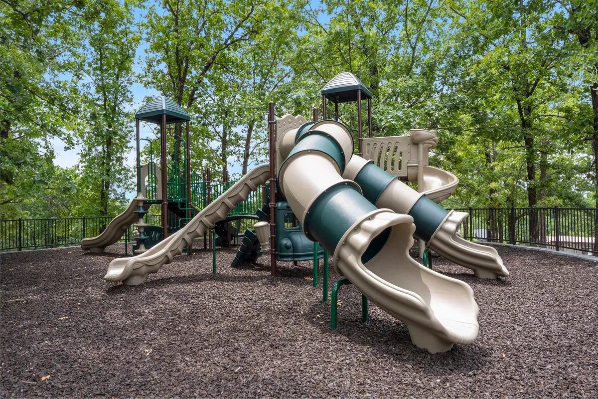 A vibrant playground with slides, situated among tall trees. The surrounding open space and greenery offer plenty of room for playful activities and family gatherings.