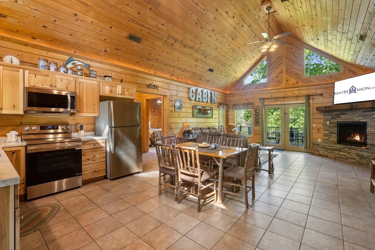 An inviting kitchen and dining space featuring rustic wooden cabinetry, a large dining table perfect for group meals, a prominent stone fireplace, and large windows that let in light and nature views. The space exudes a homey and welcoming atmosphere ideal for socializing.