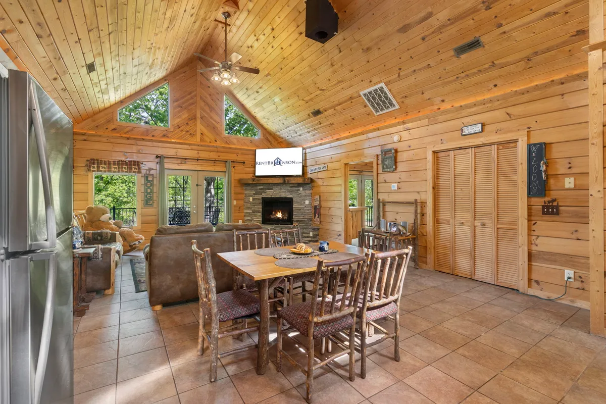 The image showcases the cabin's spacious great room, featuring a rustic wood-burning fireplace, plush couch, a sturdy dining table perfect for gatherings, and expansive windows that bathe the space in natural light. The inviting interior sets the scene for family game nights, cozy evenings by the fire, and relaxed meals together.