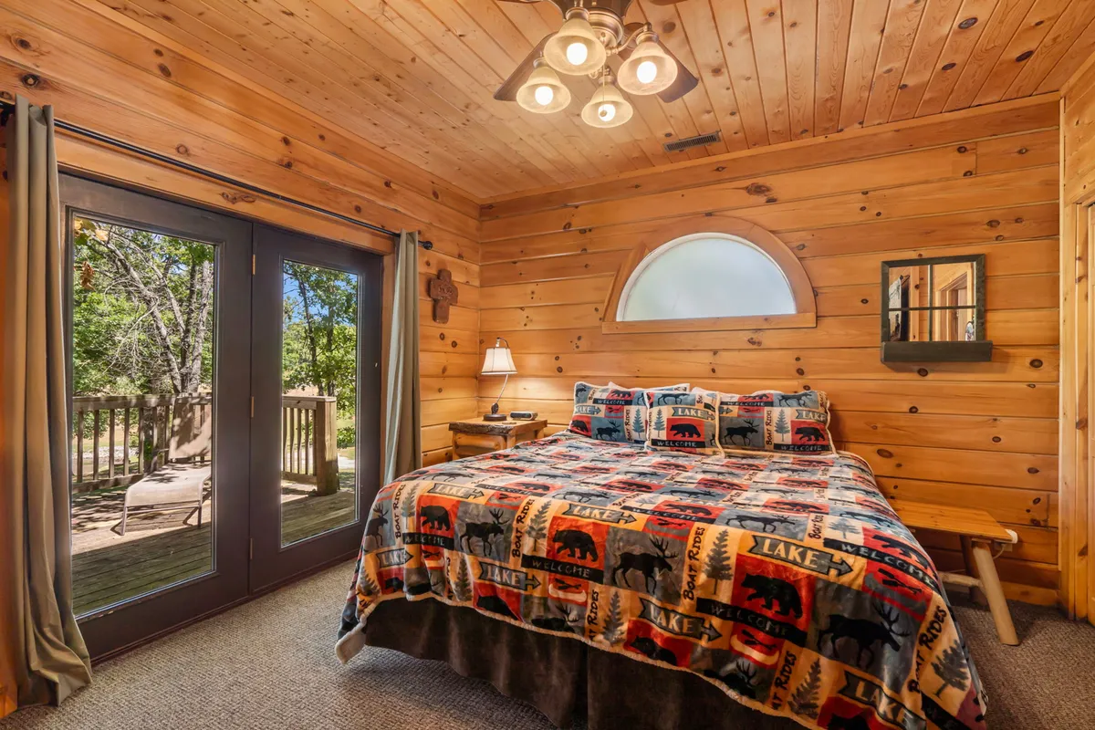 A cozy bedroom with a patterned comforter on the bed, large glass doors opening to the outside, wooden walls and ceiling, and a nightstand with a lamp. Soft natural light filters through the glass, highlighting a tranquil and inviting atmosphere.