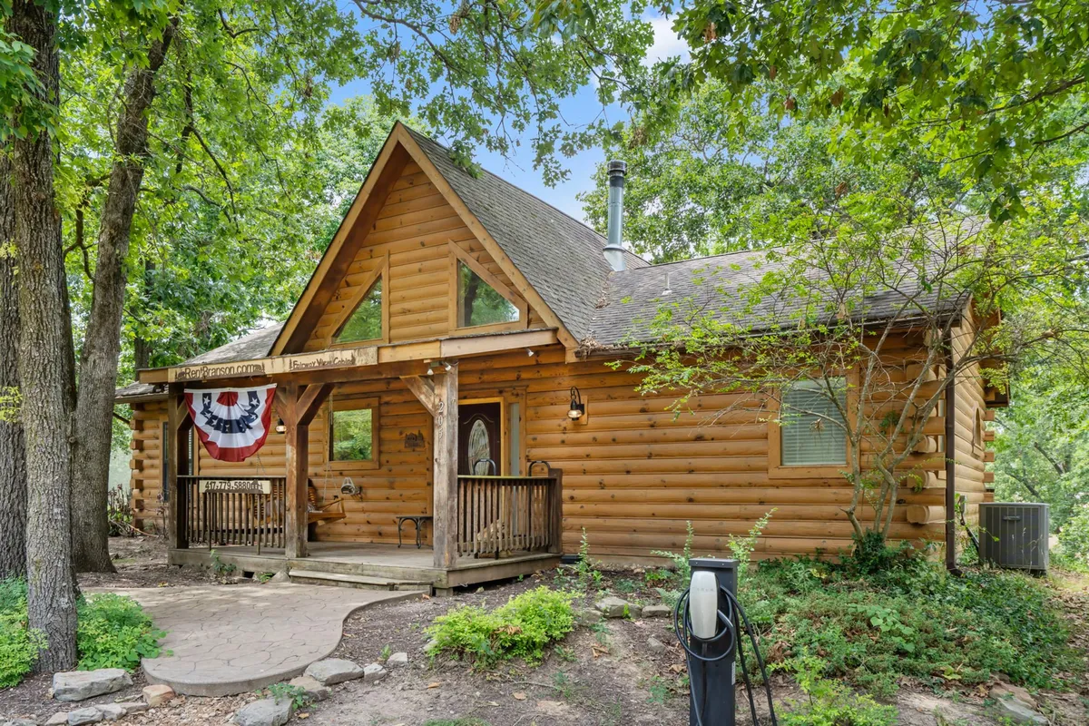 A cozy log cabin structure with a welcoming front porch draped with decorative bunting. The surrounding landscape features lush greenery and an inviting pathway, capturing a sense of connection with nature and ample space for outdoor walks.