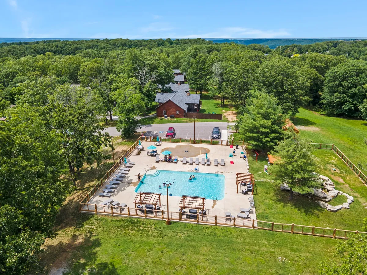 This exterior image showcases a swimming pool area with lounge chairs, cabana structures, leafy green trees, and a visible parking area in the background. The space looks modern, accessible, and thoughtfully arranged, suggesting both leisure and convenience.