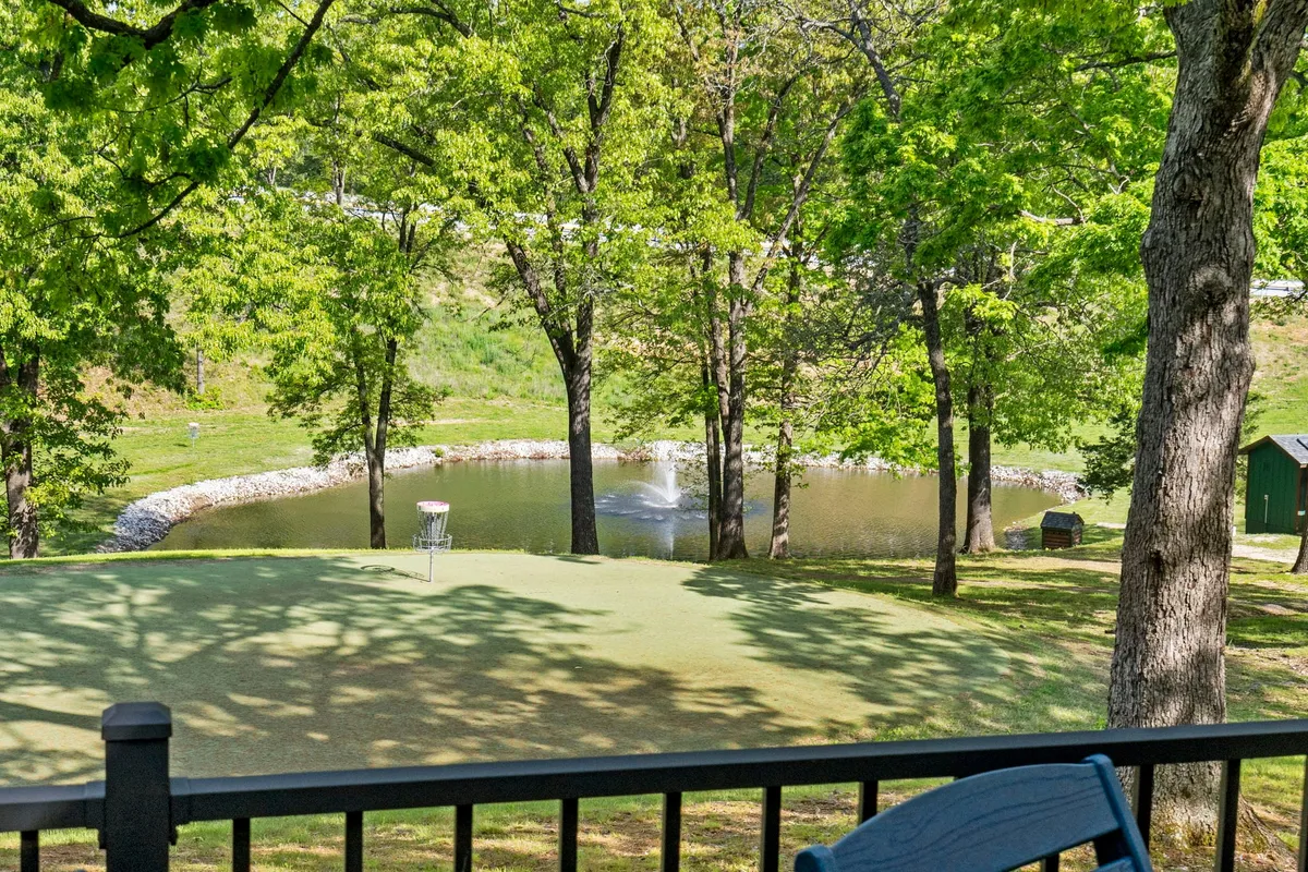 A serene pond glistens in the foreground, bordered by lush greenery and mature trees. A disc golf basket is visible near the water, suggesting recreational opportunities. The overall setting is tranquil and scenic, ideal for leisurely walks, wildlife spotting, or simply soaking in the sunset.