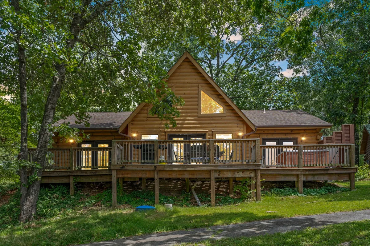 A wooden deck attached to an A-frame cabin, set among tall trees and featuring large windows. The expansive deck provides ample room for lounging and enjoying the surrounding greenery, suggesting a perfect spot to unwind after outdoor activities.