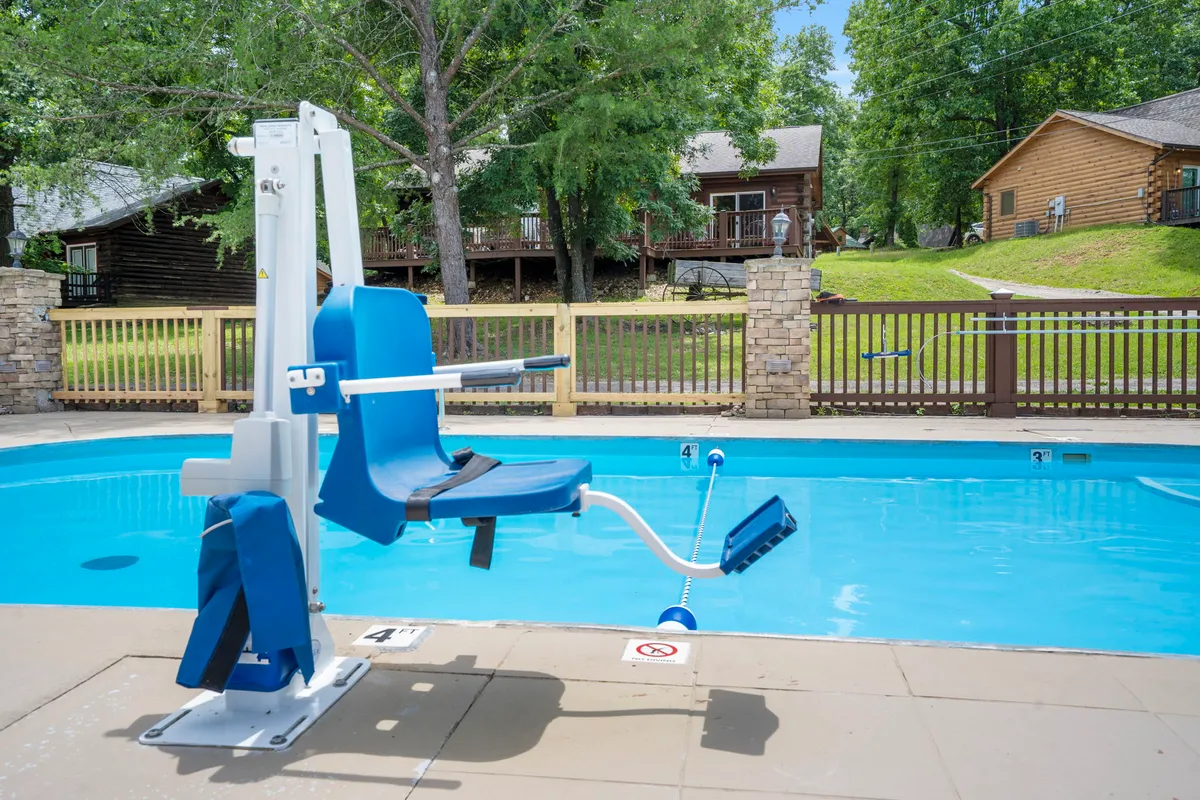 A sunny pool area surrounded by trees and cabins in the background, with a visible pool lift chair for accessibility. Families are swimming and relaxing poolside, emphasizing an inclusive and social environment.