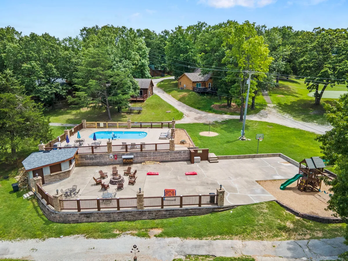 A vibrant exterior shot capturing a swimming pool at the heart of the resort, surrounded by lounge chairs, playground equipment, a basketball hoop, and cabins. Children can be seen playing, with families gathering and socializing around the communal spaces.