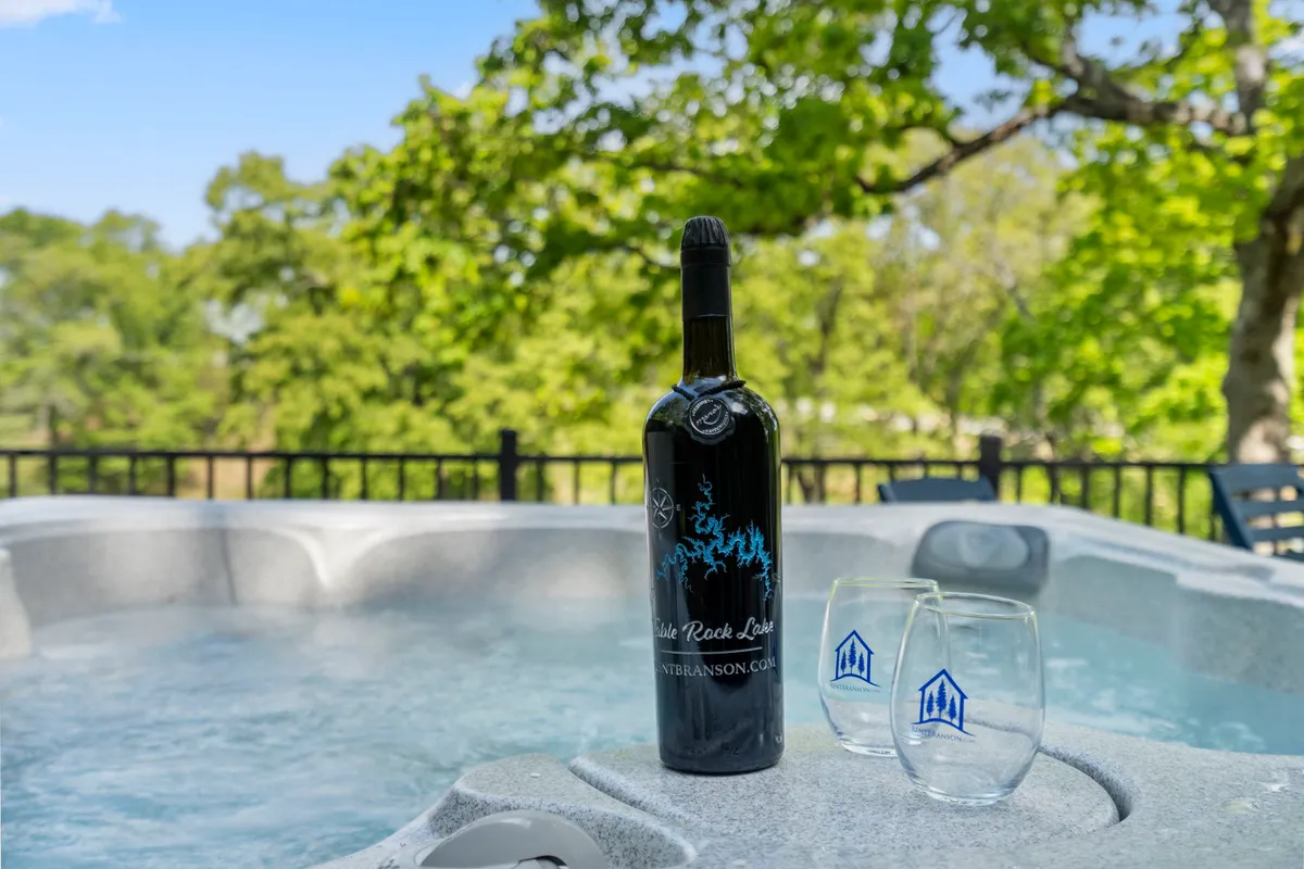This image captures a private hot tub surrounded by natural greenery, with a bottle of wine and glasses set out, suggesting a relaxing social setting. The scene evokes the idea of unwinding with friends in a tranquil outdoor environment, as the sun sets in the background.